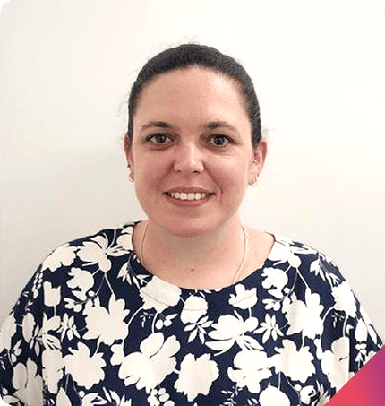 A person with dark hair tied back is smiling at the camera, embodying the warmth often found in positive behavior support plans. They are wearing a navy blue and white floral patterned top. The background is plain white, with a small pink triangle in the bottom corner of the image.