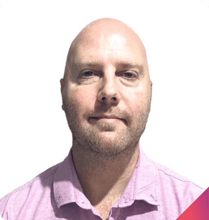 A bald man with light skin and blue eyes, wearing a light pink collared shirt, is looking directly at the camera against a plain white background, representing someone experienced in positive behaviour support plans.
