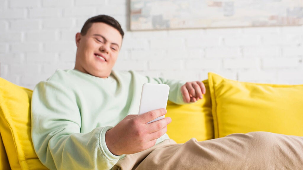 A young man with Down syndrome sits on a yellow couch, smiling while looking at his smartphone—perhaps enjoying an online behaviour therapy NDIS session. The background features a white brick wall and a light-colored painting.