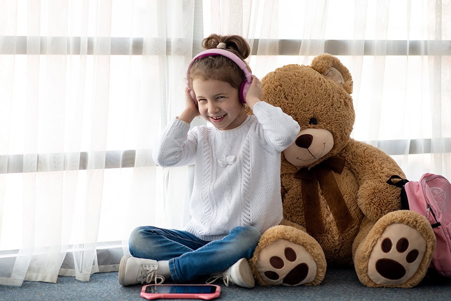 Child sitting on the floor wearing headphones beside a large teddy bear, using sound and comfort items to manage sensory needs during storms.